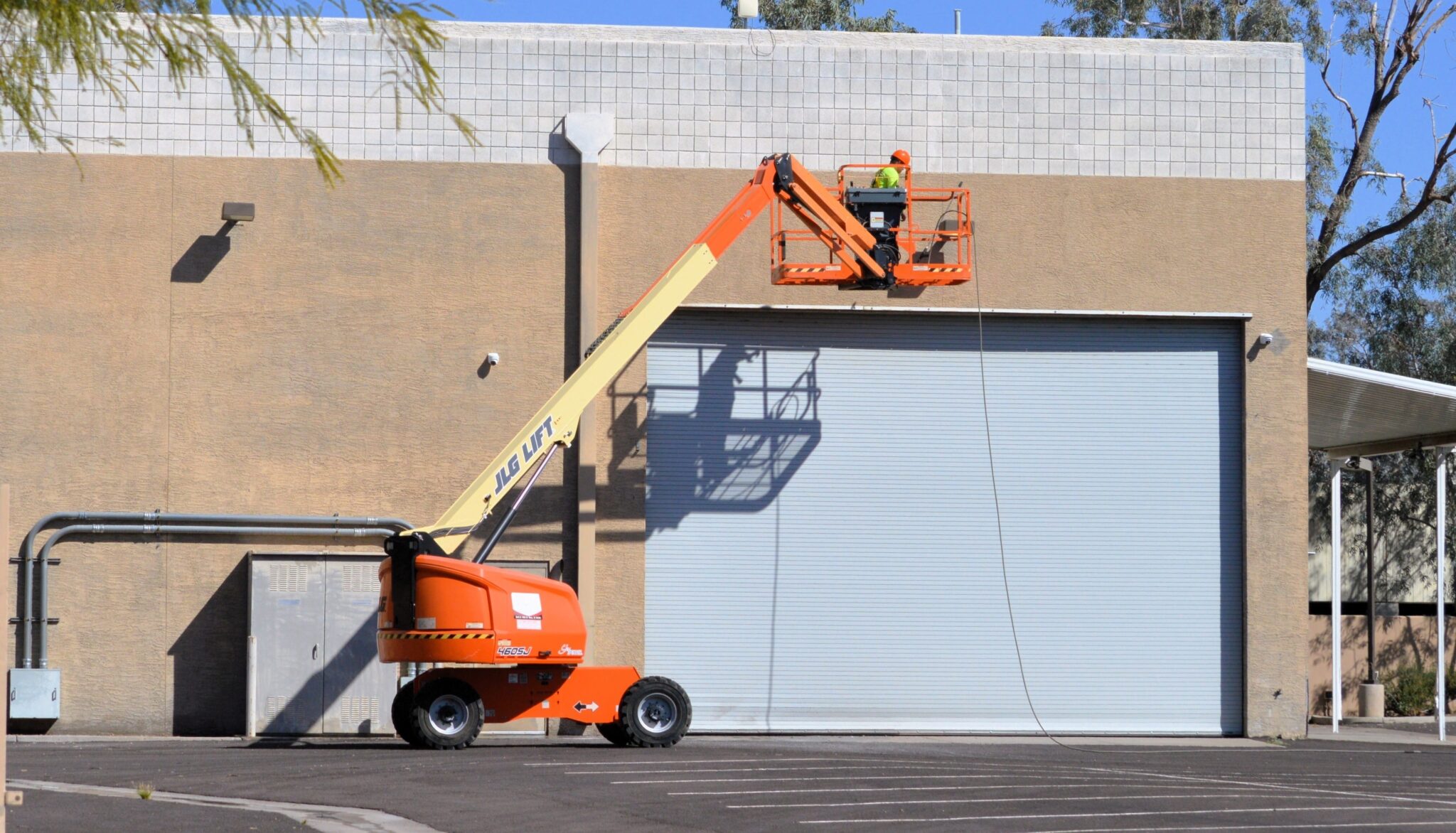A worker in a safety vest operates an elevated JLG 460SJ boom lift in front of a commercial building with a large metal roll-up door.
