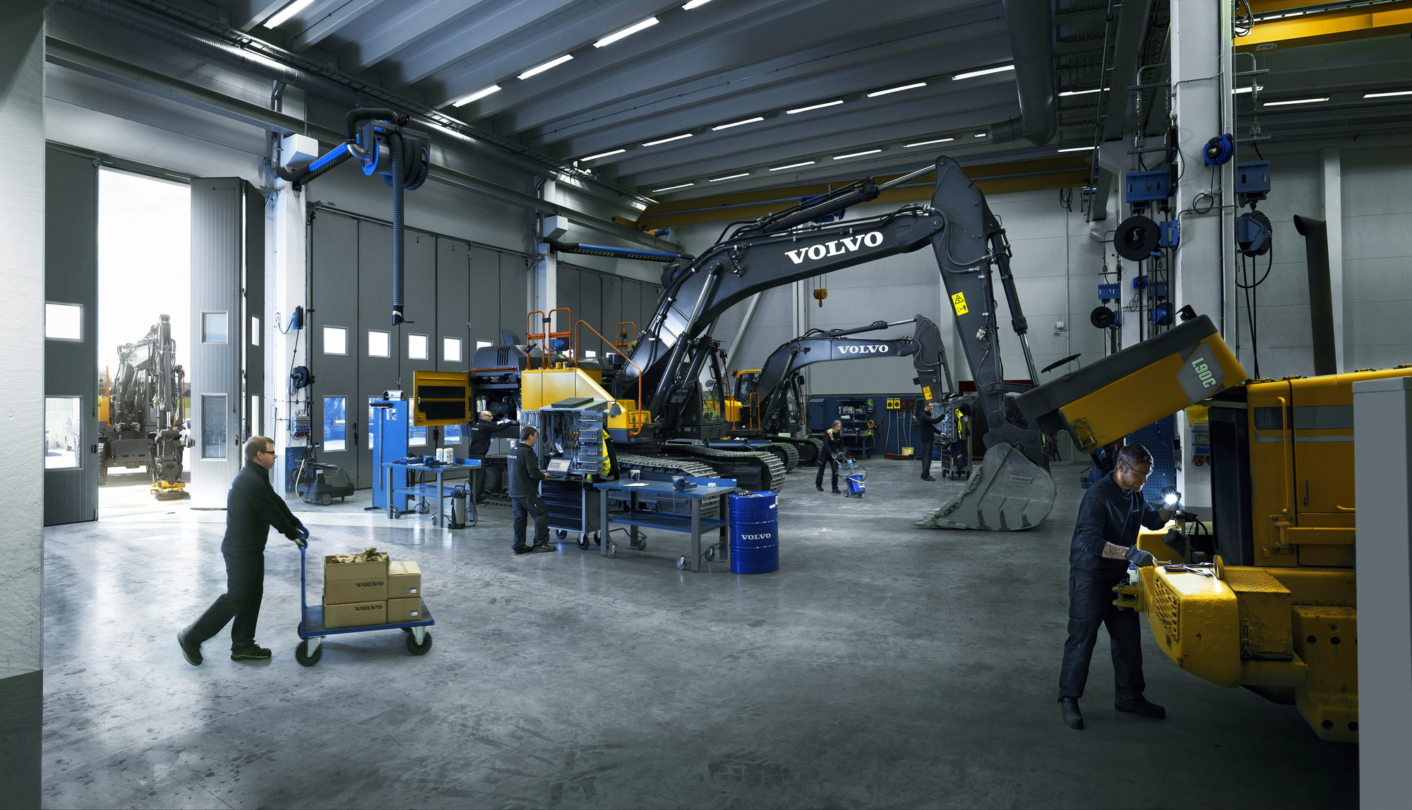 Technicians performing maintenance and repairs on large Volvo excavators and heavy machinery inside a spacious, well-lit industrial service workshop.