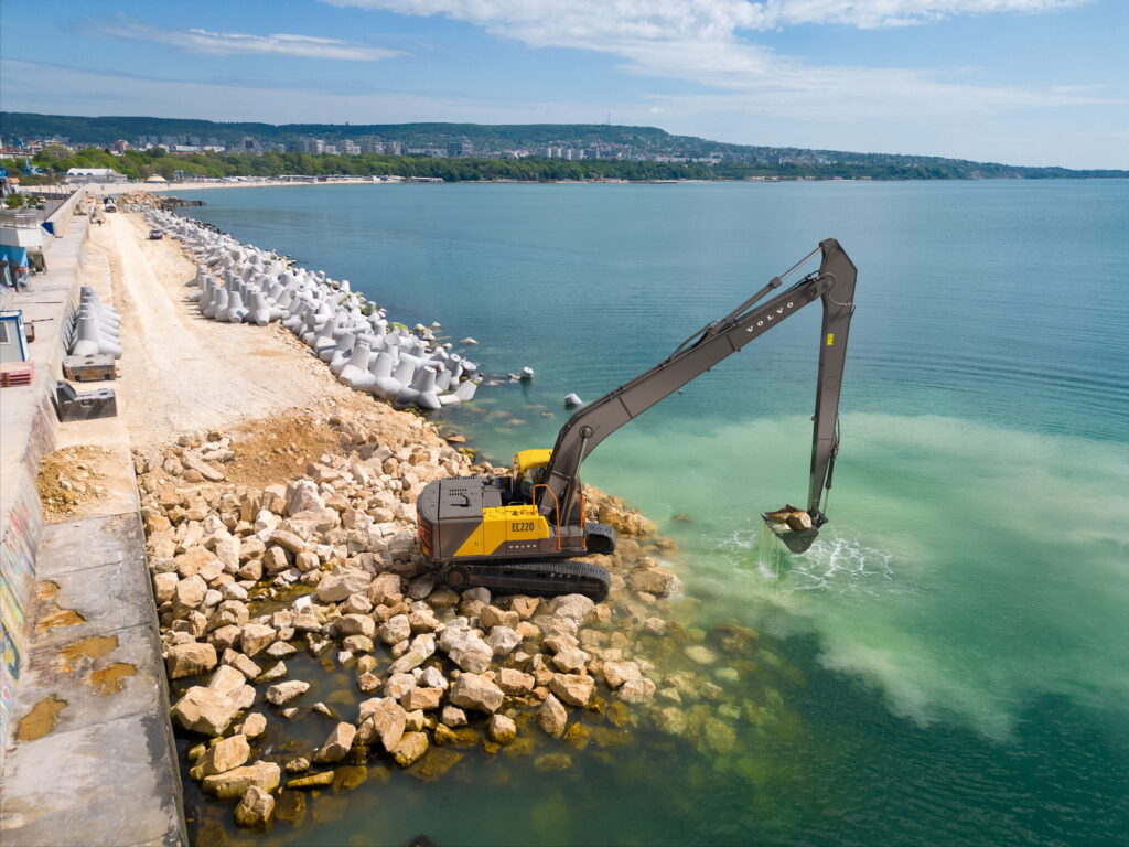 A Volvo crawler excavator with an extended long-reach arm carrying large rocks in its bucket over turquoise coastal water.
