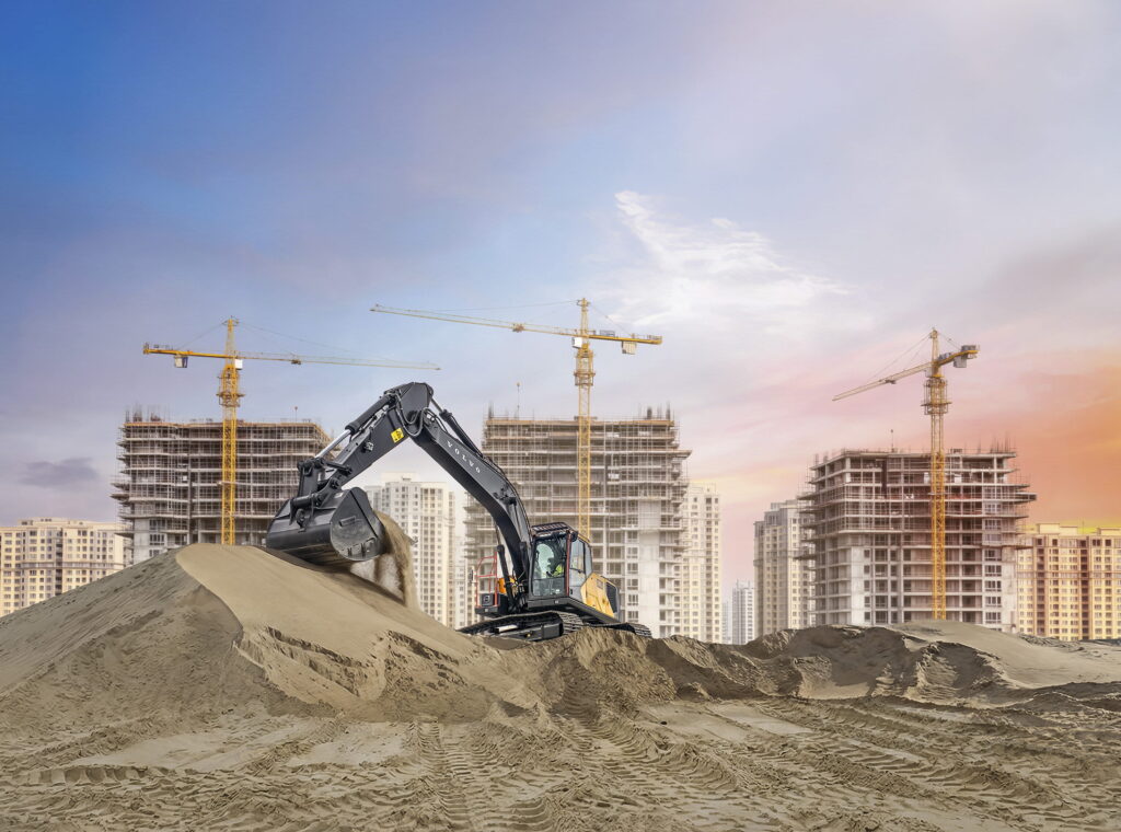 A black and yellow Volvo crawler excavator scoops sand on a large pile at a bustling high-rise construction site under a pale blue and golden sky.
