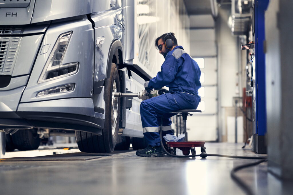 A mechanic in blue overalls and safety gear sits on a rolling stool using a pneumatic impact wrench to service the front wheel of a silver truck inside a bright workshop.