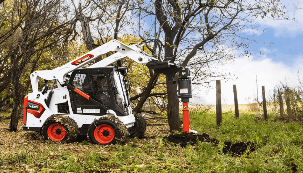 Bobcat skid steer loader drilling a hole with an auger attachment beside a tree in a grassy field